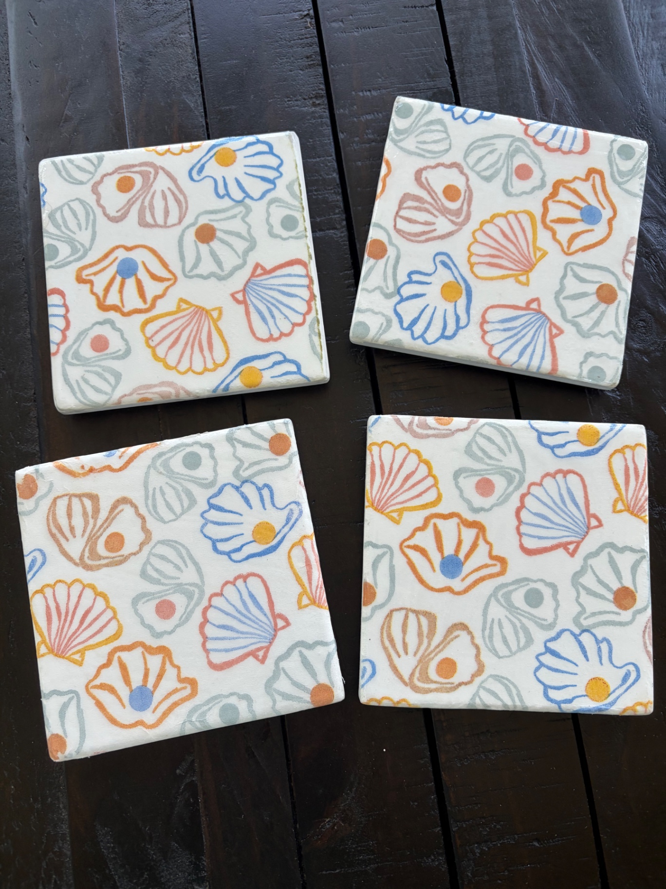 Set of four square ceramic coasters covered in a pastel pattern of shells and pearls.
