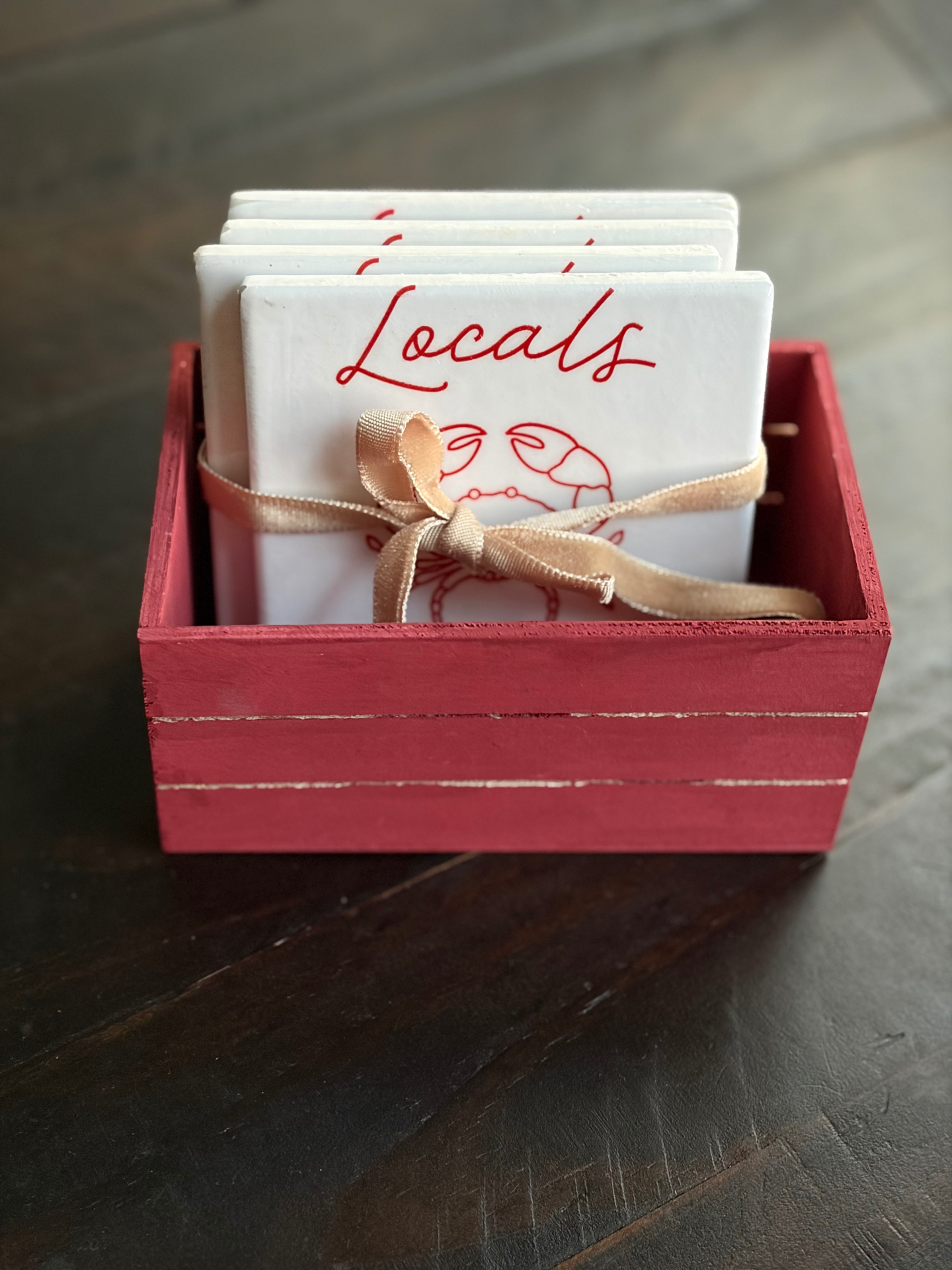 Locals Only crab coasters stacked inside a small red wooden crate, tied with a tan velvet ribbon.