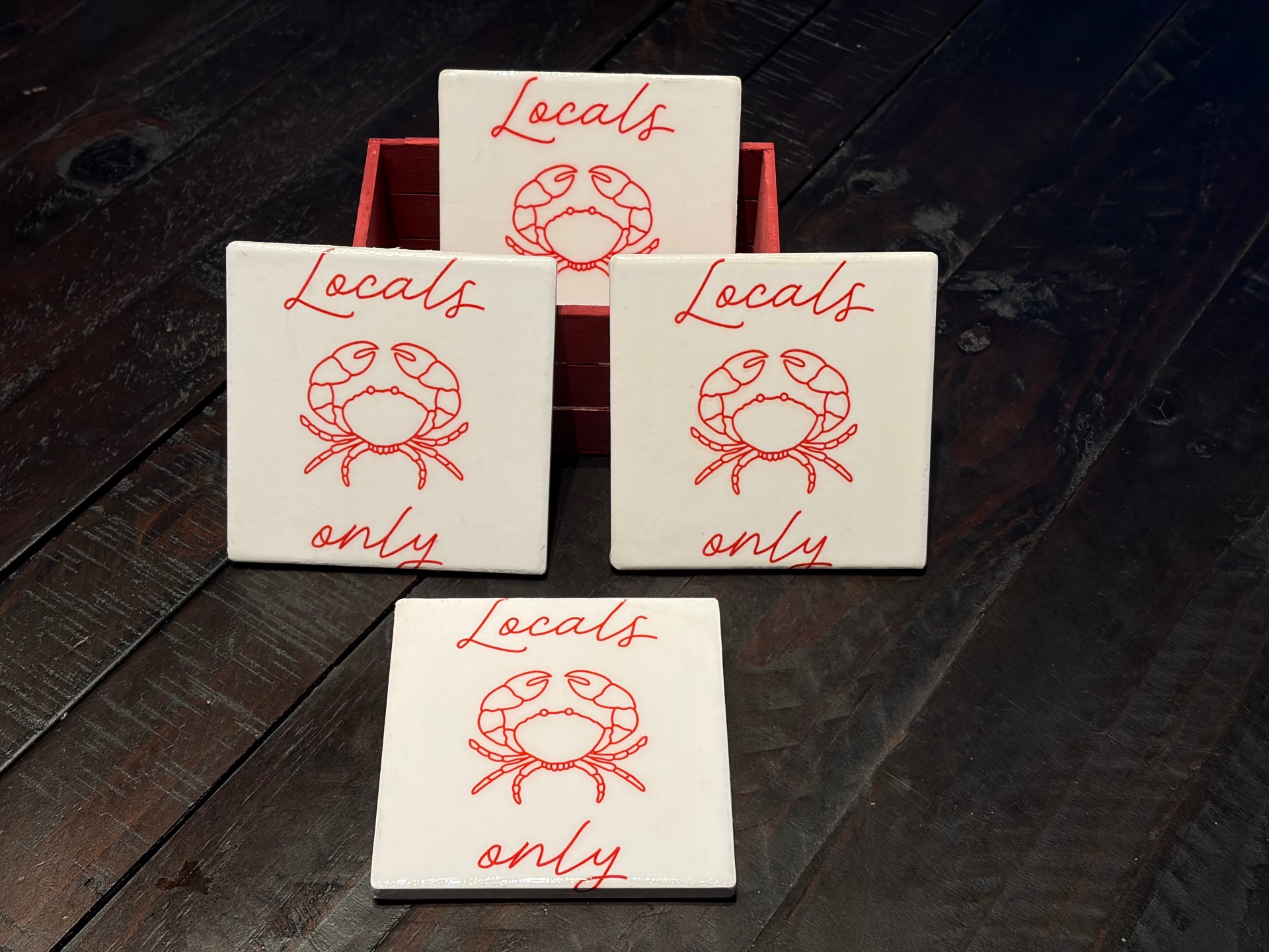 Four white square coasters, each with a red line-drawn crab and the words Locals Only in red script.
