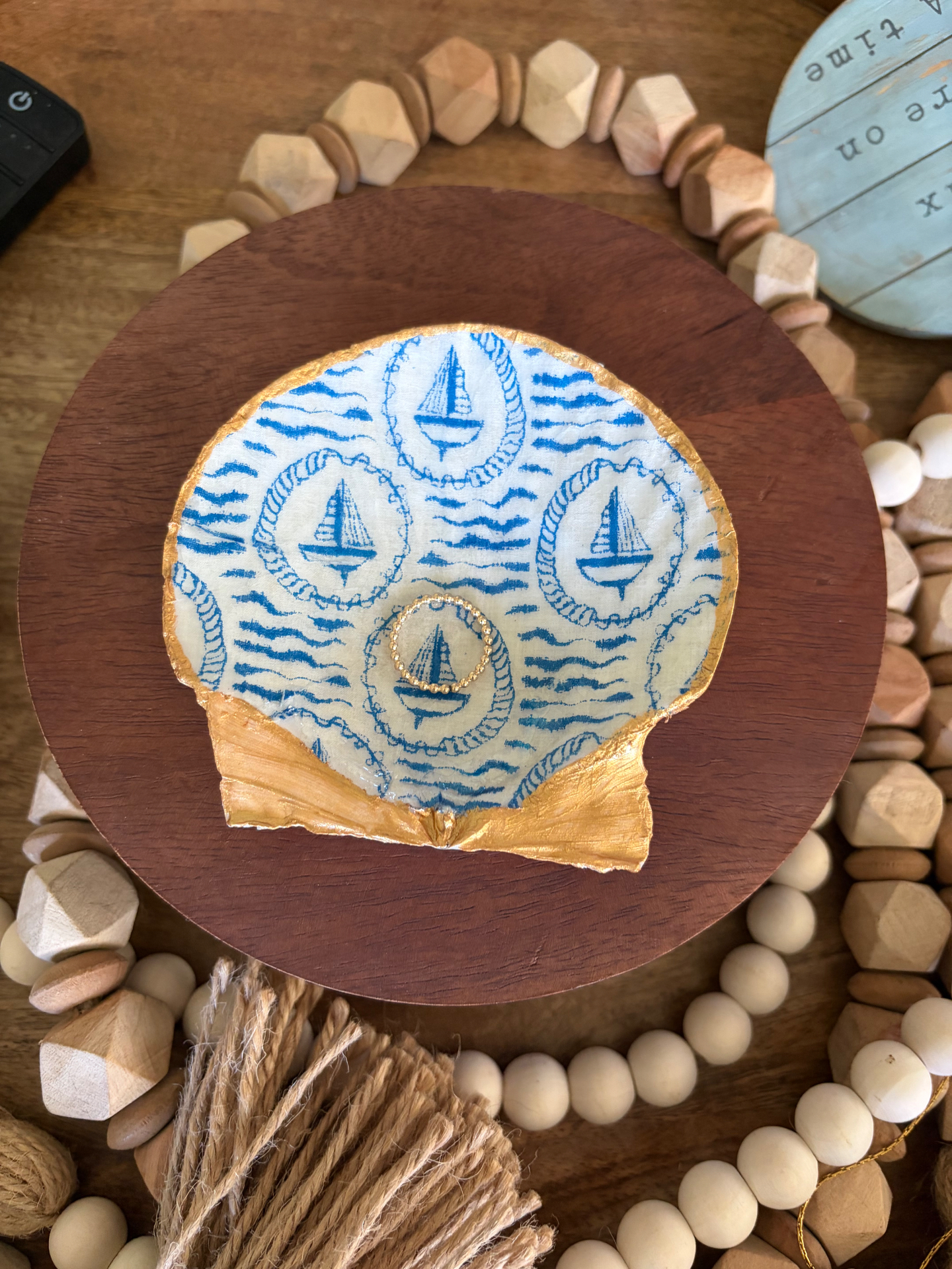 Scallop shell trinket dish decoupaged with blue sailboats and rope-framed waves, rimmed in gold and displayed on a dark wood round with wooden bead garlands (jewelry shown for styling only, not included).