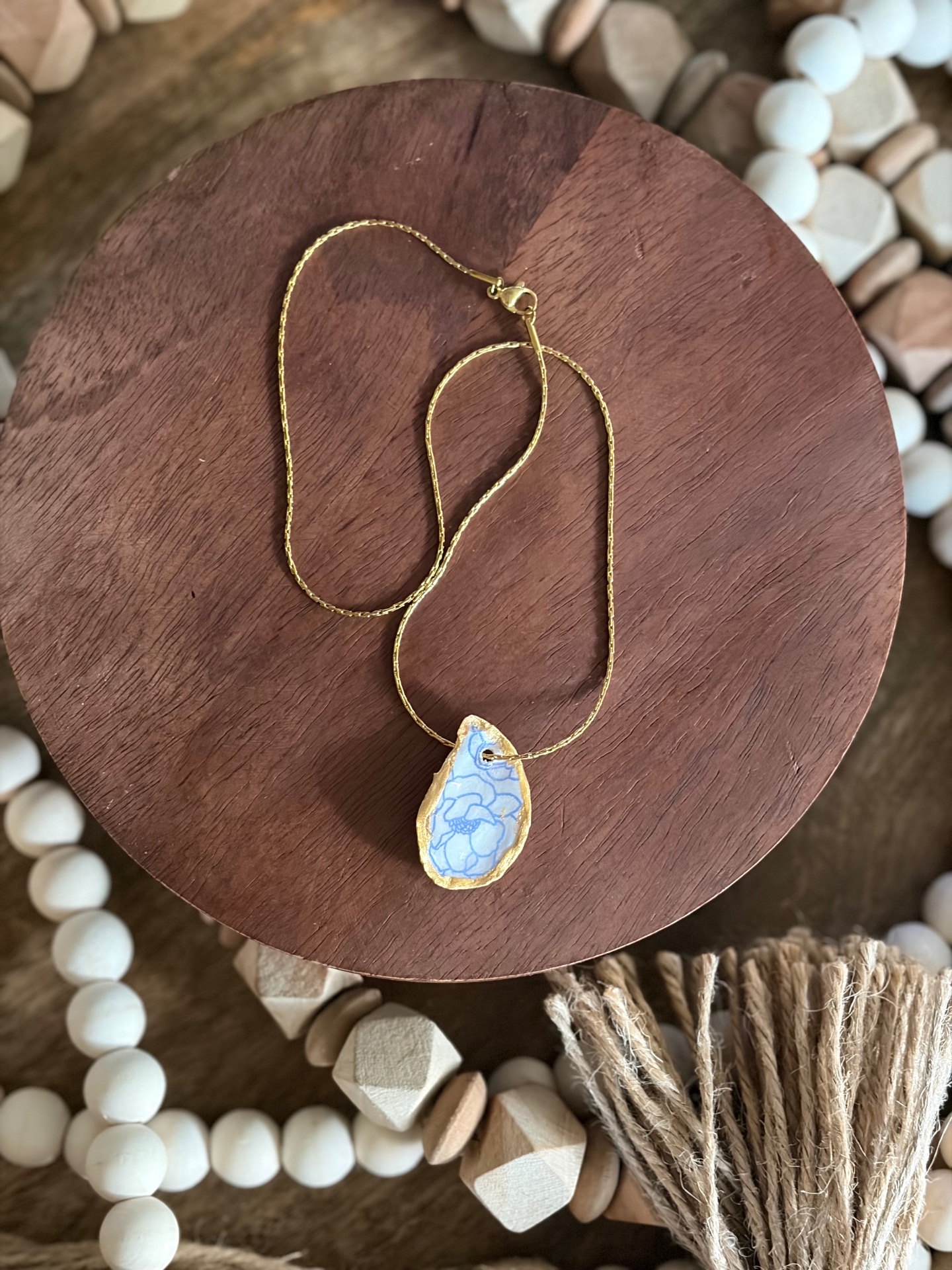 Gold chain necklace with an oyster-shell pendant hand-painted in soft blue florals and rimmed in gold, styled on a dark wood board with wooden bead garlands.