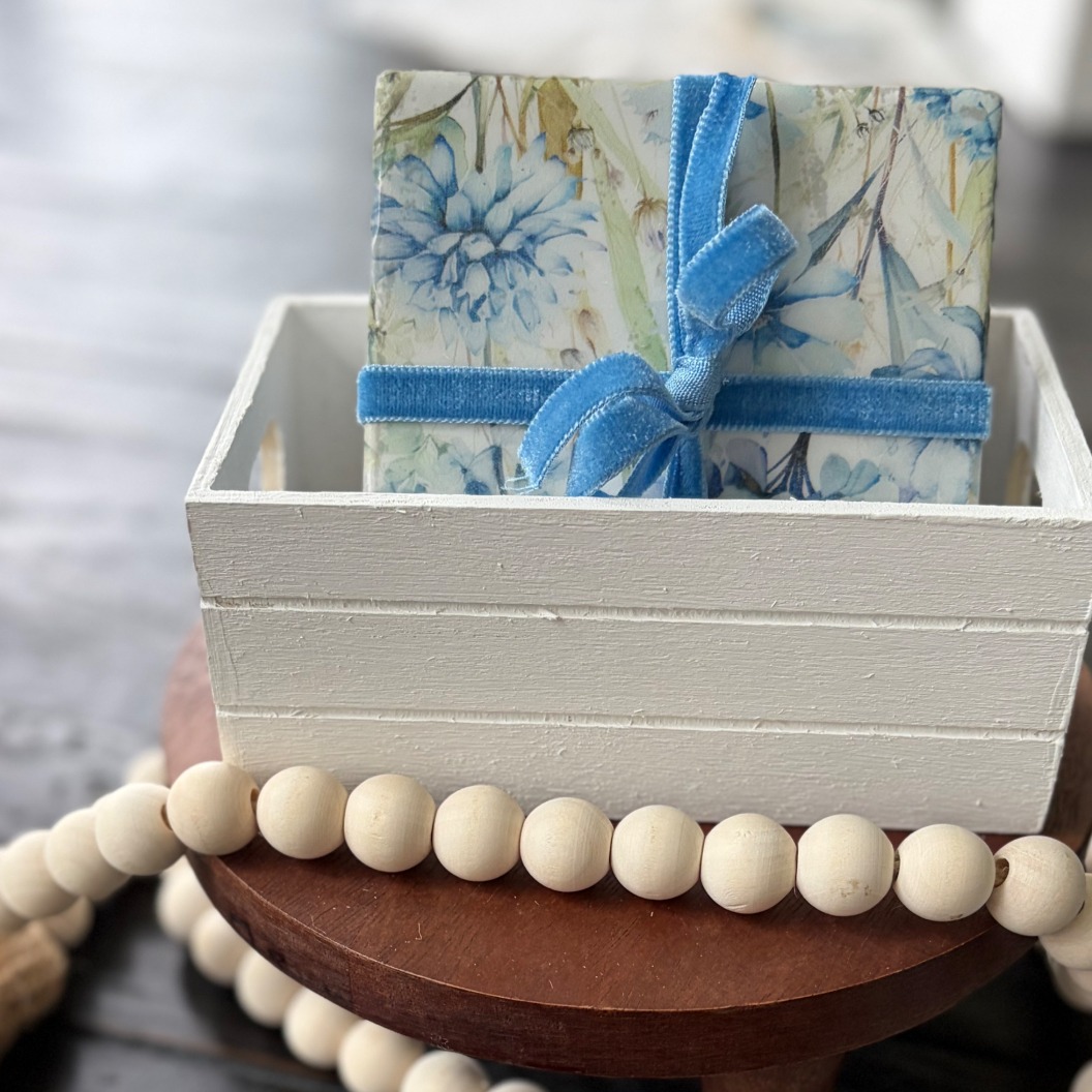 Blue hydrangea coaster set tied with blue velvet ribbon in a whitewashed wooden crate on a dark wood round.